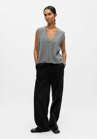 Gilet en tricot sans manches gris avec un col en V et des boutons dorés, associé à un pantalon en velours côtelé noir ample et des chaussures noires.