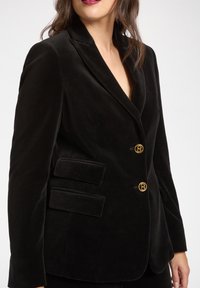 Blazer in velluto nero con collo sciallato, chiusura doppiopetto con bottoni dorati e due tasche con patta sul davanti.
