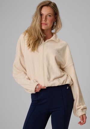 Femme aux longs cheveux blonds portant une veste polaire beige clair zippée et un pantalon bleu foncé, posant devant un fond gris uni.