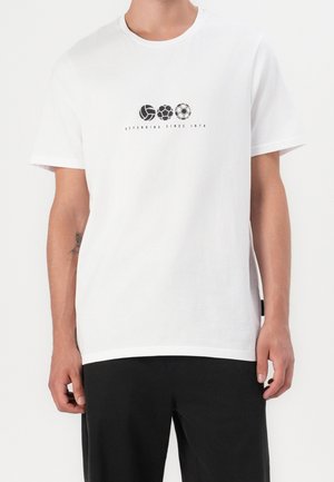 Person wearing a white t-shirt with three black sports balls and the text "DEFENDING SINCE 1874" printed across the chest.