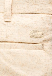 Ανοιχτό μπεζ ύφασμα με υφασμένο μοτίβο· περιλαμβάνει κεντημένο λογότυπο κροκοδείλου και επίπεδη σχεδίαση τσέπης σε ένα casual ένδυμα.