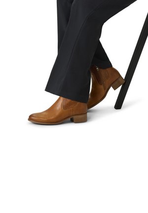 Bruine leren enkelboots met getextureerde accenten, een ronde neus, een lage blokhak en een zijrits, tegen een witte achtergrond.