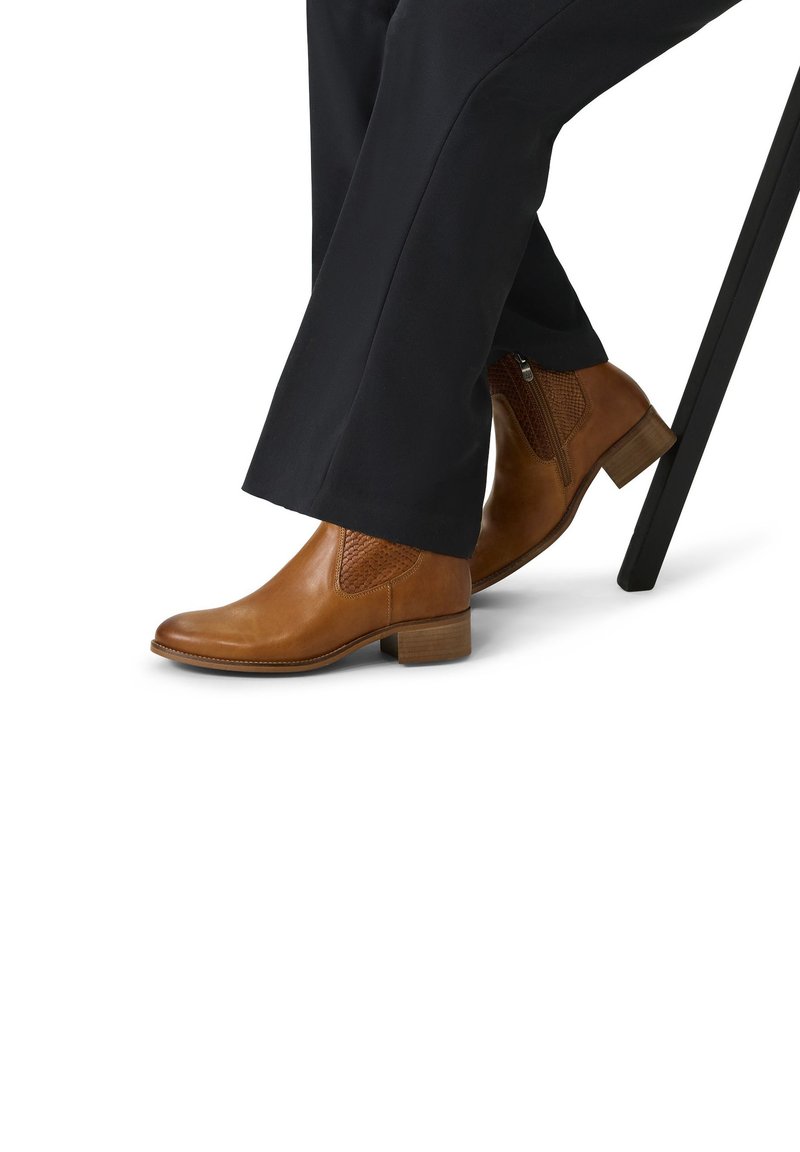 Bruine leren enkelboots met getextureerde accenten, een ronde neus, een lage blokhak en een zijrits, tegen een witte achtergrond.