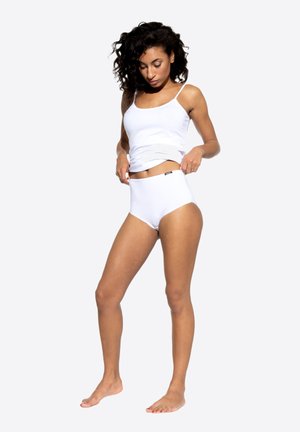 Canottiera di cotone bianca e slip a vita alta, con una texture liscia e un design semplice. La modella è in piedi a piedi nudi, mentre sistema gli slip.
