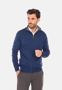 Maglione zip-up blu navy in morbido tessuto, con colletto grigio, polsini e orlo a costine. Modello in piedi, mano sollevata leggermente.