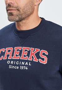 Sudadera azul marino con cuello redondo acanalado. Presenta texto en blanco y rojo "CREEKS ORIGINAL Desde 1974" en el pecho. Textura suave.