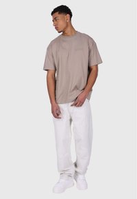 Beige short-sleeve t-shirt with a crew neckline, paired with loose white jeans and white sneakers, standing on a neutral background.