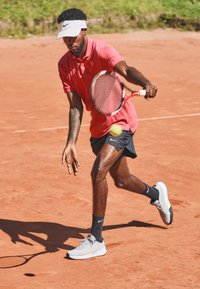 Giocatore di tennis in una polo rosa e pantaloni corti neri, con un visiera bianca e scarpe Nike grigie, pronto a colpire una palla su un campo in terra battuta.