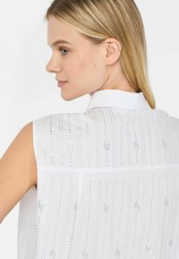 Blusa bianca senza maniche con colletto, caratterizzata da linee verticali di piccoli dettagli lucidi e una texture liscia e leggera.