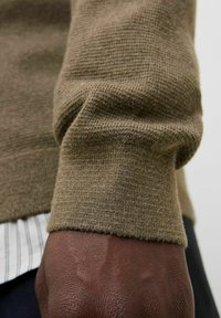 Pruunist lõngast kootud-sweateri varrukas, millel on tihe kudumine ja ribakujuline äär, mille all on valge särk, millel on peened triibud.