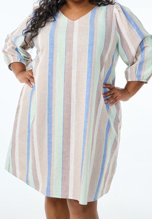 Kobieta w sukience sięgającej kolan z pionowymi pastelowymi paskami w kolorach niebieskim, zielonym, beżowym i białym, pozująca z dłońmi na biodrach.