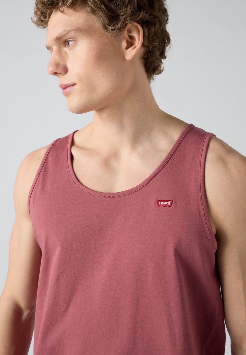 Junger Mann mit lockigem Haar, der ein ärmelloses, staubiges rosarotes Tanktop mit einem kleinen Levi's-Logo auf der Brust trägt.