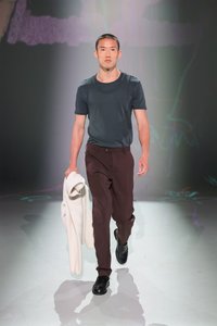 Tamno siva košulja kratkih rukava, maroon hlače, crne cipele. Model nosi bijeli, teksturirani vanjski sloj. Mekano osvjetljenje, minimalistička pozadina.