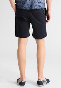 Navy blue shorts made of cotton, featuring a flat front, two back pockets, and rolled hems. Worn with casual slip-on shoes.