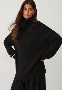 Sort strikket sweater med høj krave, brede ærmer og en afslappet pasform, parret med en plisseret sort nederdel. Teksturdetalje på stoffet er synlig.