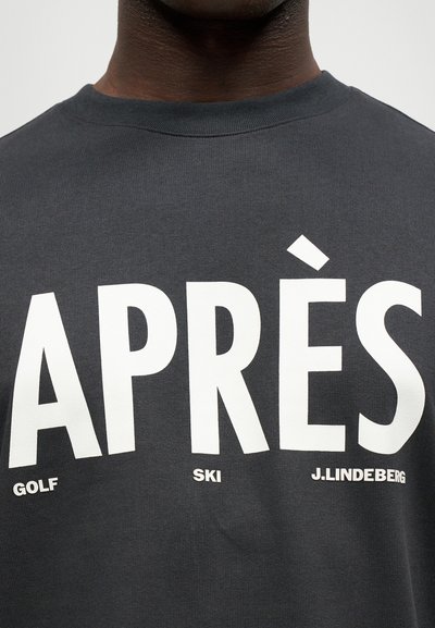 Σκούρο γκρι, μπλουζάκι με λαιμόκοψη τύπου crew που διαθέτει μεγάλο λευκό κείμενο: "APRÈS GOLF," "SKI," και "J. LINDEBERG." Κατασκευασμένο από βαμβακερό ύφασμα με ραμμένες υφές.