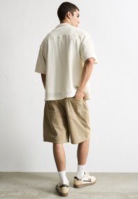 Lyse beige kortærmet skjorte med et struktureret mønster, parret med khaki knælange shorts, og hvide sokker med afslappede sneakers.