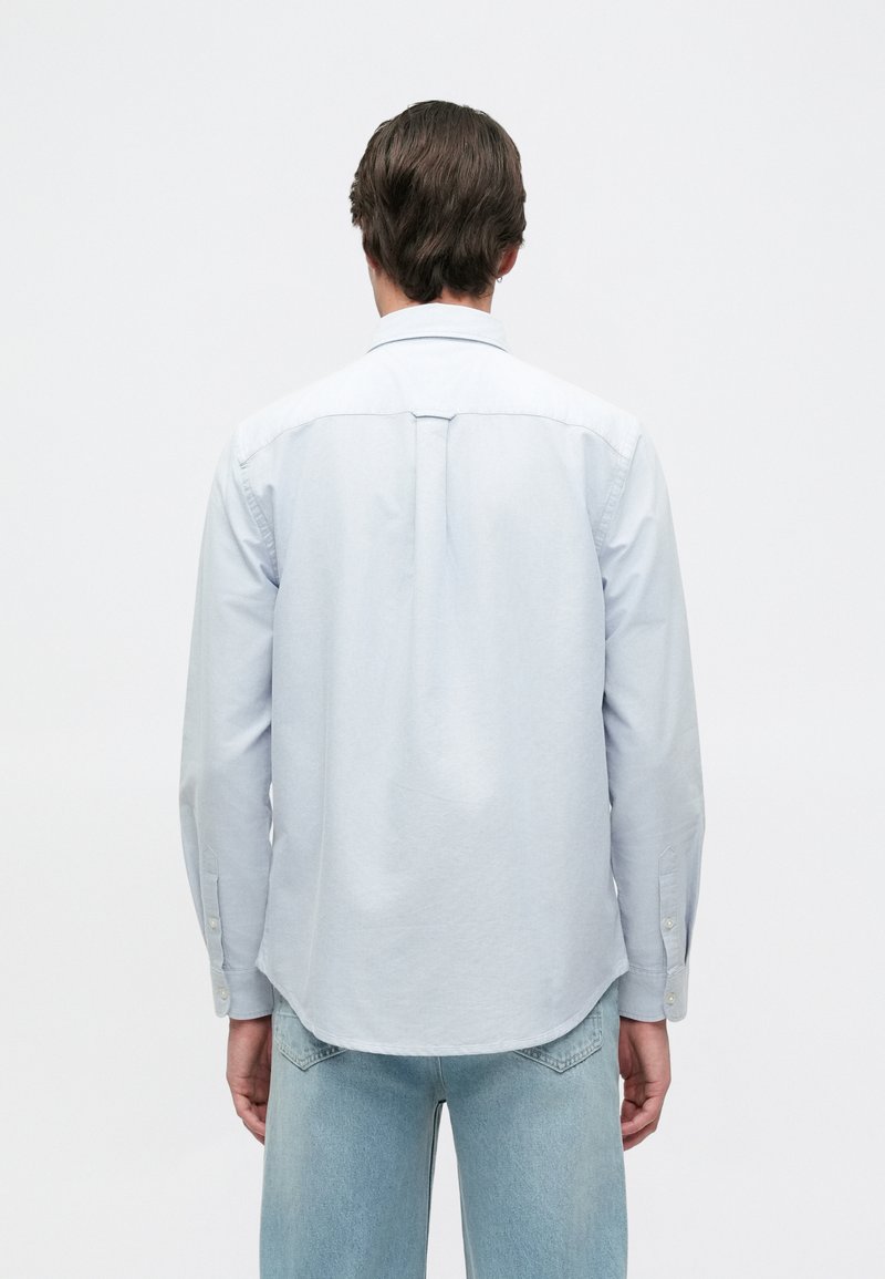 Light blue button-up shirt with long sleeves, features a back yoke, curved hem, and single-button cuffs. Smooth fabric texture.