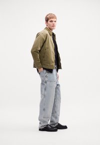Young man standing, wearing an olive green jacket, black shirt, light blue wide-leg jeans, and black shoes against a plain white background.