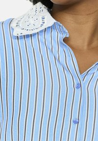 Gai zilgans krekls ar vertikālām baltām un melnām svītrām. Iekļauj baltu, izšūtu mežģīņu apkakli un zilus pogas gar priekšu.