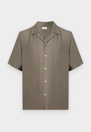 Ung mand står, iført olivengrøn kortærmet skjorte med knapper, beige bukser med vide ben, hvide sneakers og holder en brun lædertaske.