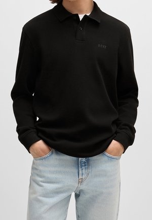 Personne portant un polo noir à manches longues avec le logo "BOSS" sur la poitrine, associé à un jean bleu clair délavé, mains dans les poches.
