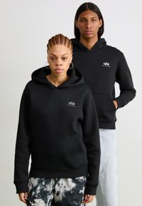 Hoodie noir en tissu doux, avec une poche avant, des poignets côtelés et un petit logo sur la poitrine. Les modèles portent des tenues décontractées.