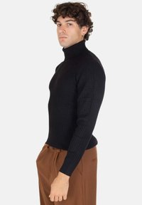 Maglione a collo alto nero a coste realizzato in tessuto morbido, dalla forma aderente, superficie testurizzata e maniche lunghe. Abbinato a pantaloni marroni.