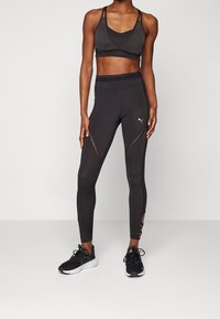 Černá sportovní podprsenka a legíny s průsvitnými panely, těsně přiléhavý design a boční výřezy. Obsahuje logo na legínách a šněrovací detaily.