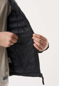 Mains tenant une veste matelassée noire ouverte montrant sa doublure intérieure lisse et une poche intérieure sur un fond uni.