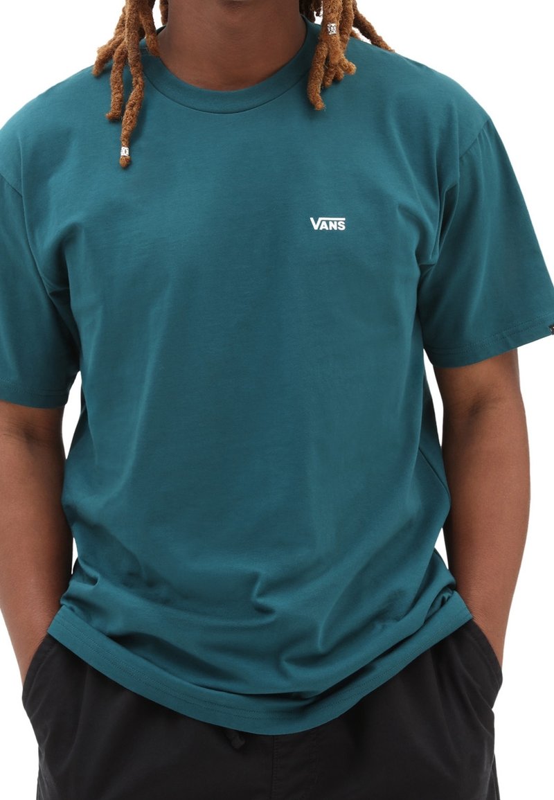 Hombre con rastas vestido con una camiseta teal de Vans y con las manos en los bolsillos de unos pantalones cortos negros, recortado del cuello hacia abajo.