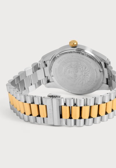 Bracelet de montre en acier inoxydable et métal doré avec une boucle gravée de "Versace" et un fond de montre rond affichant un logo Méduse.