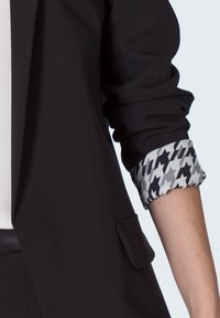 Rękaw czarnego żakietu podwinięty, ukazujący podszewkę w czarno-biały wzór w zęby psa, noszony na białej koszuli.