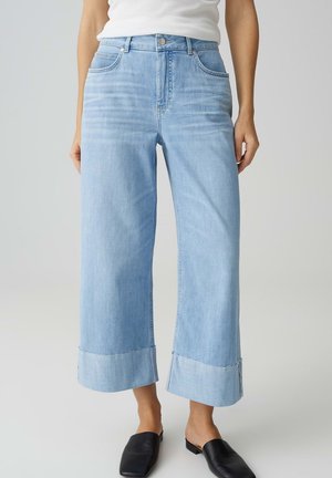 Frau trägt hellblaue, weit geschnittene, gekürzte Jeans mit rohem Saum, weißes Oberteil und schwarze Slip-On-Schuhe, steht vor einem neutralen Hintergrund.