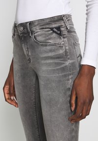 Acercamiento de una persona que lleva puestos unos jeans Replay gris desgastado con una camiseta blanca de manga larga, mostrando el bolsillo frontal y los pasadores del cinturón.