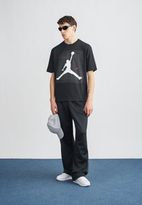 Μαύρο μπλουζάκι με λευκό γραφικό λογότυπο Jordan, συνδυασμένο με μαύρο παντελόνι φαρδιάς γραμμής και λευκά παπούτσια. Αξεσουάρ περιλαμβάνουν γκρι καπέλο και μαύρα γυαλιά ηλίου.