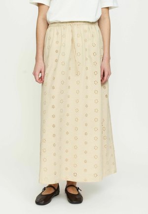 Person trägt einen beigen, knöchellangen Rock mit floralen Lochstickereien, ein weißes Oberteil und schwarze Mary Jane Schuhe und steht vor einem einfarbigen Hintergrund.