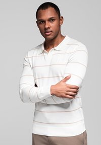 Muž s krátkými vlasy, oblečený v bílém dlouhém polo tričku s béžovými horizontálními pruhy, s překříženýma rukama, na světle šedém pozadí.