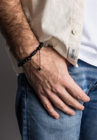 Brățară din mărgele negre cu șnururi ajustabile și mărgele decorative mici; purtată pe un încheietura mâinii deasupra unor blugi albaștri și unei cămăși bej deschis.