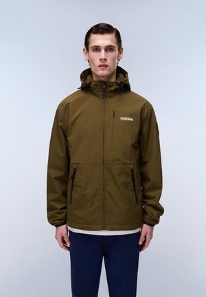 Ung mand med bagstrøget hår iført en olivengrøn hættetrøje med lynlåslommer og marineblå bukser, stående mod en ensartet lys baggrund.