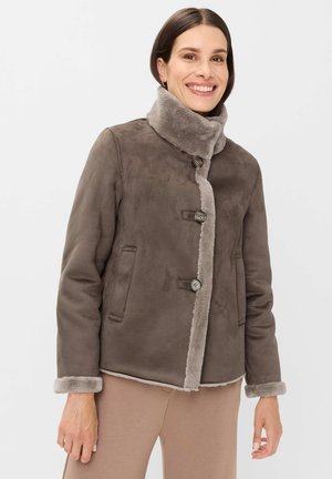 Lächelnde Frau mit brauner Lammfelljacke mit hohem Kragen, Knopfverschluss und Seitentaschen, kombiniert mit beigen Hosen.