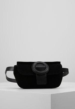 Zwarte suède heuptas met een brede riem en een grote ronde gesp aan de voorkant, geplaatst op een wit voetstuk tegen een grijze achtergrond.