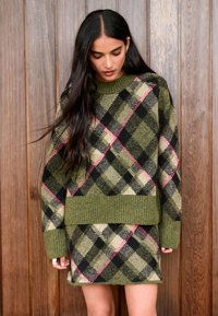 Maglione verde a quadri in maglia con maniche lunghe e orlo a coste, abbinato a una gonna coordinata con accenti rosa e neri. Sfondo in legno.