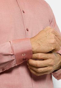 Hellrosa Langarmhemd aus strukturierter Stoffqualität mit dunklen Knöpfen und kontrastierenden Manschettendetails, gehalten von Händen, die die Ärmelsäume anpassen.