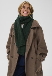 Bufanda verde texturizada alrededor del cuello, llevada con un abrigo de lana marrón que tiene grandes botones y un corte entallado. Fondo liso.