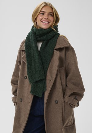 Groene textuur sjaal om de nek gedrapeerd, gedragen met een bruine wollen jas met grote knopen en een getailleerde snit. Effen achtergrond.