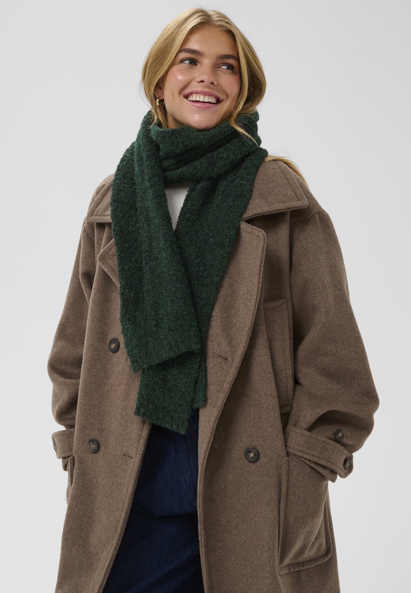 Bufanda verde texturizada alrededor del cuello, llevada con un abrigo de lana marrón que tiene grandes botones y un corte entallado. Fondo liso.