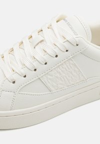 White leather sneaker with textured branding, flat white laces, and a low profile. Features a smooth finish and subtle stitching details.