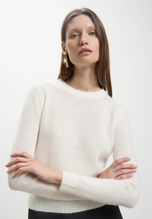 Maglione a maglia color crema con collo rotondo, polsini a coste e una consistenza morbida. Il modello posa con le braccia incrociate, indossando orecchini d'oro.