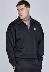 Čierna mikina na zips s stojatým golierom, vyrobená z hladkej látky, s malým vyšitým logom na hrudi a vo voľnom strihu.